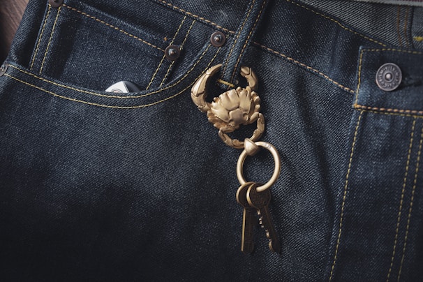 Dark denim jeans featuring a prominent brass keychain shaped like a crab, with a set of keys attached. The fabric's texture and stitching details are visible, highlighting the contrast between the dark denim and the golden keychain.