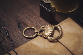 A gold-colored crab keychain lies on top of a brown leather notebook on a wooden surface. Nearby, there is a metallic lighter and a black beaded bracelet, contributing to a vintage aesthetic.