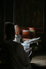Two individuals are seated in a dimly lit room, both engrossed in reading newspapers. The setting is calm with natural light filtering into the space, creating a warm and cozy atmosphere. A stack of colorful books or magazines is visible in the background, adding a touch of color to the scene.
