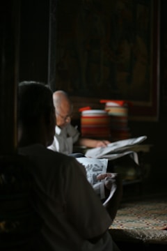 Two individuals are seated in a dimly lit room, both engrossed in reading newspapers. The setting is calm with natural light filtering into the space, creating a warm and cozy atmosphere. A stack of colorful books or magazines is visible in the background, adding a touch of color to the scene.