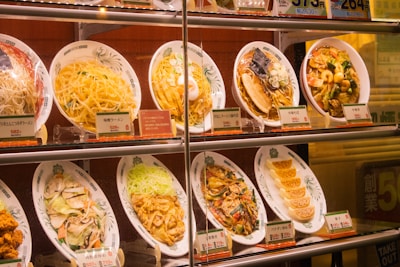 A display case with several plastic food replicas showcasing various dishes, including noodles and dumplings. Each dish is accompanied by a small card with prices and labels in Japanese, indicating typical menu options at a restaurant.