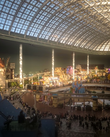 A large indoor amusement park with a high glass ceiling, featuring a monorail track, various colorful attractions, and a crowd of people. The space is spacious and brightly lit, with a whimsical and lively atmosphere.