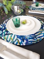 A sophisticated dining table setting featuring a decorative placemat with a blue and green leaf pattern, set with a white ceramic plate and a small green glass. A slice of lime rests in a shallow dish on the plate. The setting includes a white napkin with textured silverware. Lush foliage and purple flowers are arranged at one end of the table, and an elegant glass sits nearby.