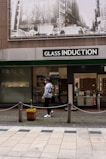 A person walks past a storefront with large signage that reads 'Glass Induction'. Above the store, there is a grayscale image of a busy city street with buildings and traffic. The sidewalk is patterned with light and dark tiles, and a potted plant with flowers is placed near a chain barrier.