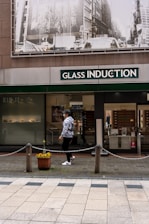 A person walks past a storefront with large signage that reads 'Glass Induction'. Above the store, there is a grayscale image of a busy city street with buildings and traffic. The sidewalk is patterned with light and dark tiles, and a potted plant with flowers is placed near a chain barrier.