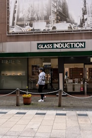 A person walks past a storefront with large signage that reads 'Glass Induction'. Above the store, there is a grayscale image of a busy city street with buildings and traffic. The sidewalk is patterned with light and dark tiles, and a potted plant with flowers is placed near a chain barrier.