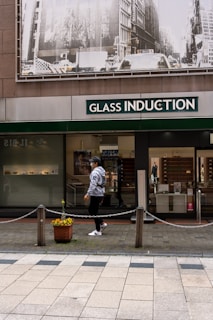 A person walks past a storefront with large signage that reads 'Glass Induction'. Above the store, there is a grayscale image of a busy city street with buildings and traffic. The sidewalk is patterned with light and dark tiles, and a potted plant with flowers is placed near a chain barrier.