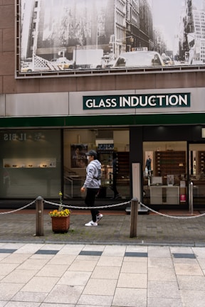 A person walks past a storefront with large signage that reads 'Glass Induction'. Above the store, there is a grayscale image of a busy city street with buildings and traffic. The sidewalk is patterned with light and dark tiles, and a potted plant with flowers is placed near a chain barrier.