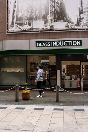 A person walks past a storefront with large signage that reads 'Glass Induction'. Above the store, there is a grayscale image of a busy city street with buildings and traffic. The sidewalk is patterned with light and dark tiles, and a potted plant with flowers is placed near a chain barrier.
