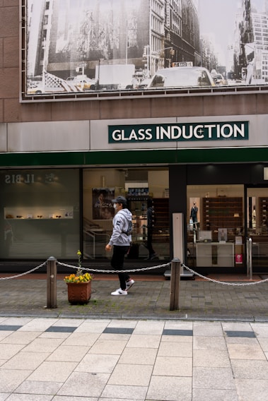 A person walks past a storefront with large signage that reads 'Glass Induction'. Above the store, there is a grayscale image of a busy city street with buildings and traffic. The sidewalk is patterned with light and dark tiles, and a potted plant with flowers is placed near a chain barrier.