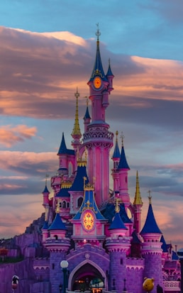 A vibrant, fairy-tale castle with multiple colorful spires stands majestically against a backdrop of dramatic, dusky clouds. The architecture features a blend of pink, blue, and gold hues, evoking a sense of magic and wonder. The castle is illuminated with warm lights, enhancing its enchanting appearance.