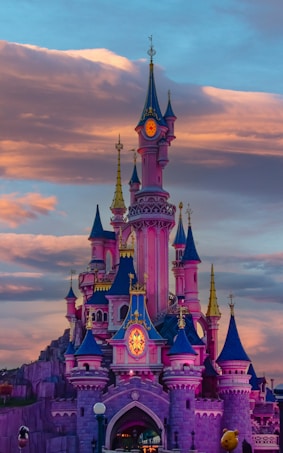 A vibrant, fairy-tale castle with multiple colorful spires stands majestically against a backdrop of dramatic, dusky clouds. The architecture features a blend of pink, blue, and gold hues, evoking a sense of magic and wonder. The castle is illuminated with warm lights, enhancing its enchanting appearance.