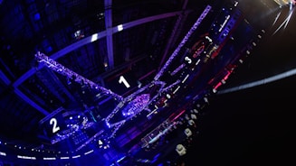 A large indoor arena with colorful and vibrant lighting, featuring a center stage surrounded by rigging and structures. Large numbered screens are visible, indicating marked sections from one to five. Various pieces of equipment and people are scattered around the venue.