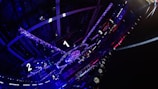 A large indoor arena with colorful and vibrant lighting, featuring a center stage surrounded by rigging and structures. Large numbered screens are visible, indicating marked sections from one to five. Various pieces of equipment and people are scattered around the venue.