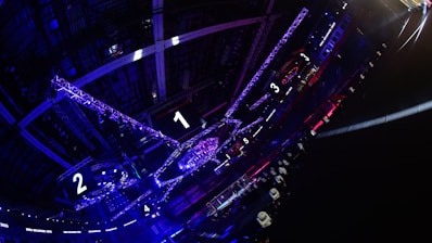 A large indoor arena with colorful and vibrant lighting, featuring a center stage surrounded by rigging and structures. Large numbered screens are visible, indicating marked sections from one to five. Various pieces of equipment and people are scattered around the venue.