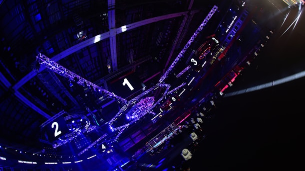 A large indoor arena with colorful and vibrant lighting, featuring a center stage surrounded by rigging and structures. Large numbered screens are visible, indicating marked sections from one to five. Various pieces of equipment and people are scattered around the venue.