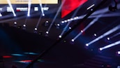 An array of bright stage lights beams across a dark arena, creating a dynamic atmosphere with a large screen displaying various logos and text. A camera crane is visible in the foreground, suggesting a professional event setup.
