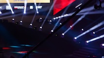 An array of bright stage lights beams across a dark arena, creating a dynamic atmosphere with a large screen displaying various logos and text. A camera crane is visible in the foreground, suggesting a professional event setup.