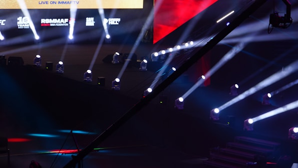 An array of bright stage lights beams across a dark arena, creating a dynamic atmosphere with a large screen displaying various logos and text. A camera crane is visible in the foreground, suggesting a professional event setup.