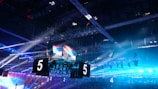A large indoor arena is equipped with advanced lighting and audio systems. Suspended above the space are numerous digital displays showcasing numbers. The environment is filled with blue illumination and spotlights creating a vibrant atmosphere.
