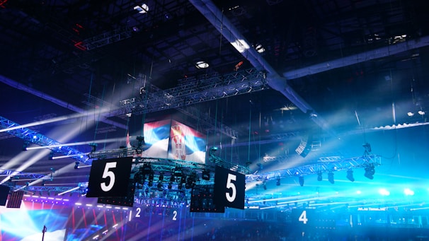 A large indoor arena is equipped with advanced lighting and audio systems. Suspended above the space are numerous digital displays showcasing numbers. The environment is filled with blue illumination and spotlights creating a vibrant atmosphere.