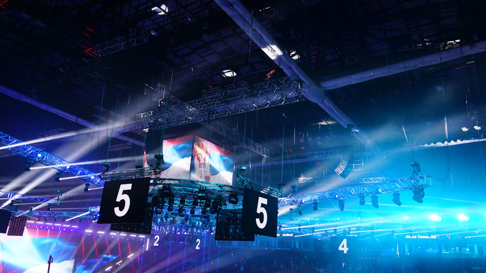 A large indoor arena is equipped with advanced lighting and audio systems. Suspended above the space are numerous digital displays showcasing numbers. The environment is filled with blue illumination and spotlights creating a vibrant atmosphere.