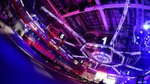 An indoor arena with vibrant purple and blue lighting, featuring large numbered screens suspended from the ceiling. The venue has a high ceiling with intricate metal truss structures and hanging lights, creating a futuristic and energetic atmosphere.