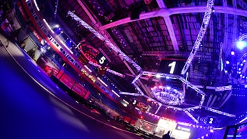 An indoor arena with vibrant purple and blue lighting, featuring large numbered screens suspended from the ceiling. The venue has a high ceiling with intricate metal truss structures and hanging lights, creating a futuristic and energetic atmosphere.