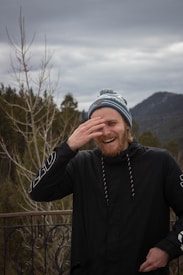 A person is laughing with one hand covering part of their face, standing on a balcony with a mountainous background. The sky is overcast, and trees without leaves are visible.