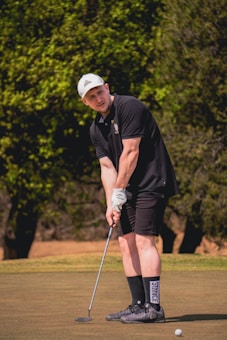 A person wearing a black polo shirt, black shorts, and a white cap is standing on a golf course, holding a putter and seemingly preparing to take a golf shot. The background features green trees under bright sunlight.