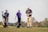 Three men are on a golf course. One man is preparing to hit the golf ball with a club, while the other two stand nearby. A golf bag with various clubs is present, and they are surrounded by lush greenery and a clear sky.