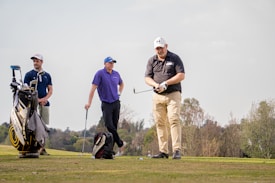 Three men are on a golf course. One man is preparing to hit the golf ball with a club, while the other two stand nearby. A golf bag with various clubs is present, and they are surrounded by lush greenery and a clear sky.
