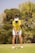 A person wearing a yellow shirt, white visor, and white shorts is preparing to hit a golf ball on a lush green golf course. The background features tall trees and foliage indicative of a sunny day.