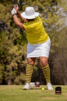 A person is playing golf, wearing a yellow shirt, white hat, and striped yellow and black socks. The individual is in a swinging stance, focusing on hitting a small yellow golf ball on a green course with trees in the background.