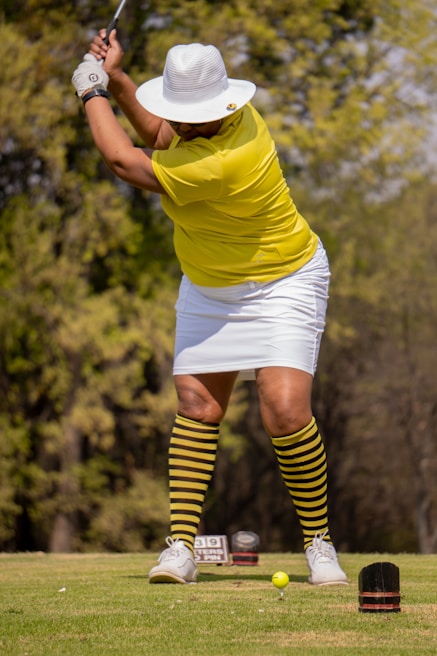 A person is playing golf, wearing a yellow shirt, white hat, and striped yellow and black socks. The individual is in a swinging stance, focusing on hitting a small yellow golf ball on a green course with trees in the background.