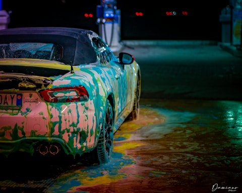 A car at a wash station covered in colorful soap suds, creating a vibrant and artistic pattern on its surface. The car's bodywork reflects a range of colors under the station's lights, and the ground is covered with swirling soap in various hues.