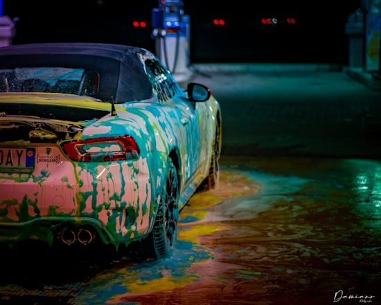 A car at a wash station covered in colorful soap suds, creating a vibrant and artistic pattern on its surface. The car's bodywork reflects a range of colors under the station's lights, and the ground is covered with swirling soap in various hues.