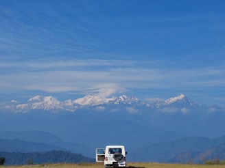 A white SUV is parked on a grassy field with its back door open, against a backdrop of majestic snow-capped mountains under a clear blue sky. The serene landscape captures the vastness of nature and the sense of adventure.