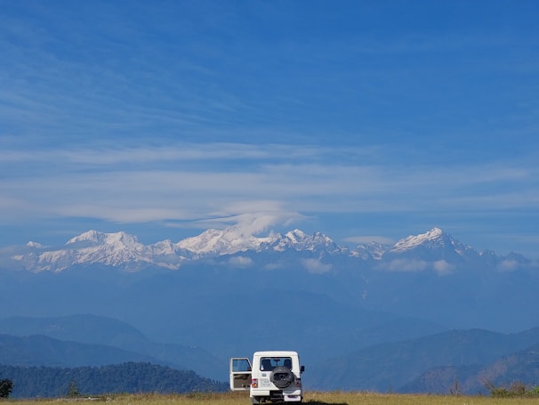 A white SUV is parked on a grassy field with its back door open, against a backdrop of majestic snow-capped mountains under a clear blue sky. The serene landscape captures the vastness of nature and the sense of adventure.