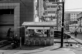 A small burger stand sits on the corner of a busy street in an urban environment. The kiosk is covered by a roof and displays bright signage with the word 'BURGER' and various cartoon characters. Surrounding the stand are tall buildings, electric wires, and a closed storefront. A motorcycle is parked nearby, while pedestrians and cars move in the background. The scene is captured in black and white, adding a nostalgic, gritty feel.