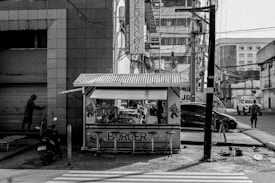 A small burger stand sits on the corner of a busy street in an urban environment. The kiosk is covered by a roof and displays bright signage with the word 'BURGER' and various cartoon characters. Surrounding the stand are tall buildings, electric wires, and a closed storefront. A motorcycle is parked nearby, while pedestrians and cars move in the background. The scene is captured in black and white, adding a nostalgic, gritty feel.