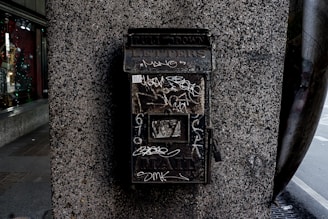 A weathered metal mailbox is mounted on a textured stone wall. The mailbox has graffiti scribbled on its surface and the word 'LETTERS' is partially visible at the top. It appears old and slightly rusty, suggesting it has been in place for a long time. A glass window display is visible on the left, featuring various items and faint reflections.