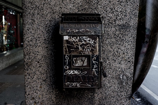 A weathered metal mailbox is mounted on a textured stone wall. The mailbox has graffiti scribbled on its surface and the word 'LETTERS' is partially visible at the top. It appears old and slightly rusty, suggesting it has been in place for a long time. A glass window display is visible on the left, featuring various items and faint reflections.