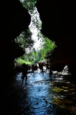 A group of people walking through a narrow, rocky canyon with a stream flowing at their feet. The silhouette of the rocks frames a bright sky above, with green foliage cascading down the sides, providing a contrast to the shadows. The scene conveys a sense of exploration and adventure.