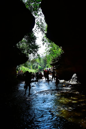 A group of people walking through a narrow, rocky canyon with a stream flowing at their feet. The silhouette of the rocks frames a bright sky above, with green foliage cascading down the sides, providing a contrast to the shadows. The scene conveys a sense of exploration and adventure.