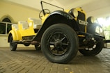 A vintage car with bright yellow bodywork and black accents is parked indoors on a stone-paved floor. The car features large round headlights, visible carriage wheels, and an open-top design with cushioned seats. A window with an arch design is in the background, along with some greenery visible through another opening.