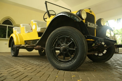A vintage car with bright yellow bodywork and black accents is parked indoors on a stone-paved floor. The car features large round headlights, visible carriage wheels, and an open-top design with cushioned seats. A window with an arch design is in the background, along with some greenery visible through another opening.