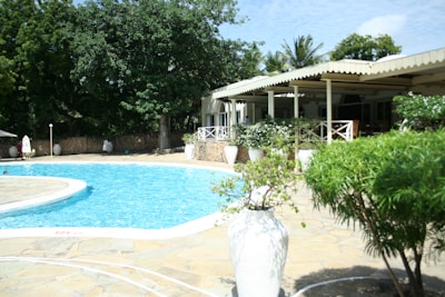 A serene outdoor setting with a large swimming pool surrounded by a stone patio. Lush green trees and shrubs provide shade and privacy. A white open-air structure with a roof offers seating and dining areas, adorned with white railings. Large white planters line the poolside, adding to the tropical and tranquil ambiance.
