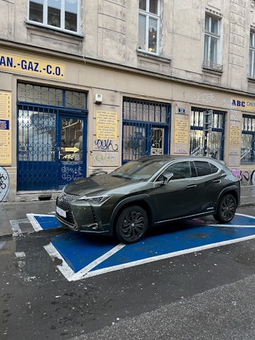 A dark-colored car is parked in a designated parking spot with blue and white marking lines, adjacent to a building. The building has beige walls and multiple signs in a foreign language displayed above blue metal gates. The walls are decorated with graffiti, and there are several windows with white frames on the upper level.
