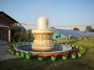 A large decorative fountain is surrounded by vibrant green and red potted plants. The area is well-maintained with a paved walkway and a building adorned with festive banners in the background. In the distance, solar panels and a mountain can be seen under a clear blue sky.